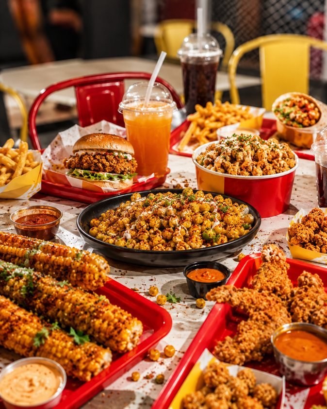 A table spread of corn ribs, crispy corn, a burger, fries and a wrap on red trays.